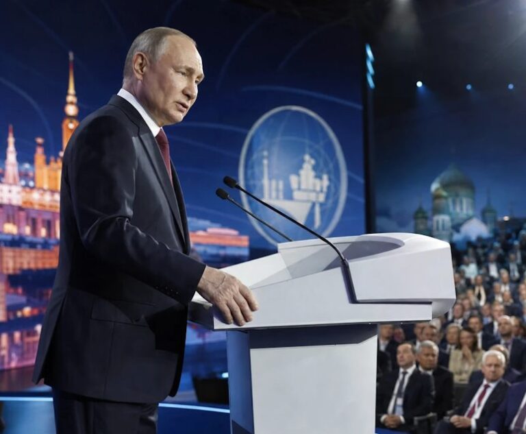 Man in a dark suit speaking at a lectern on a large stage, with a blue backdrop and a seated audience.